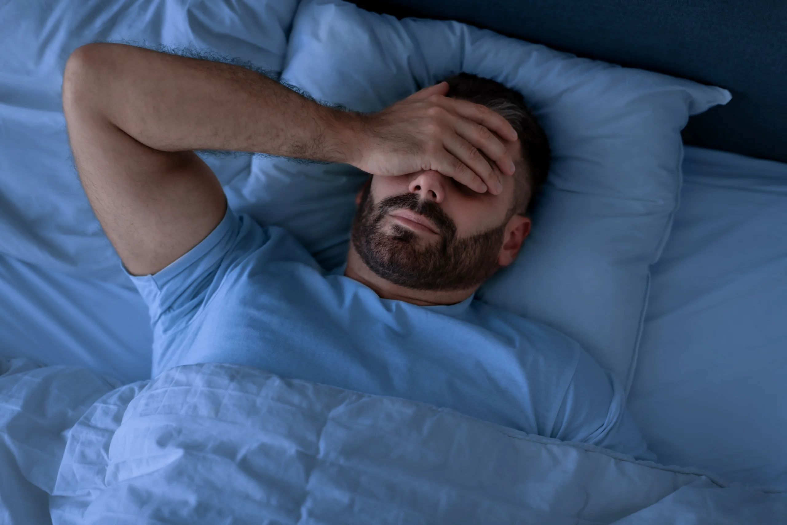 Insomnia Problem. Man Covering Face With Hand Lying Unable To Sleep In Bed At Home, Suffering From Headache And Sleeplessness In Modern Bedroom Interior. Top View, Low Light