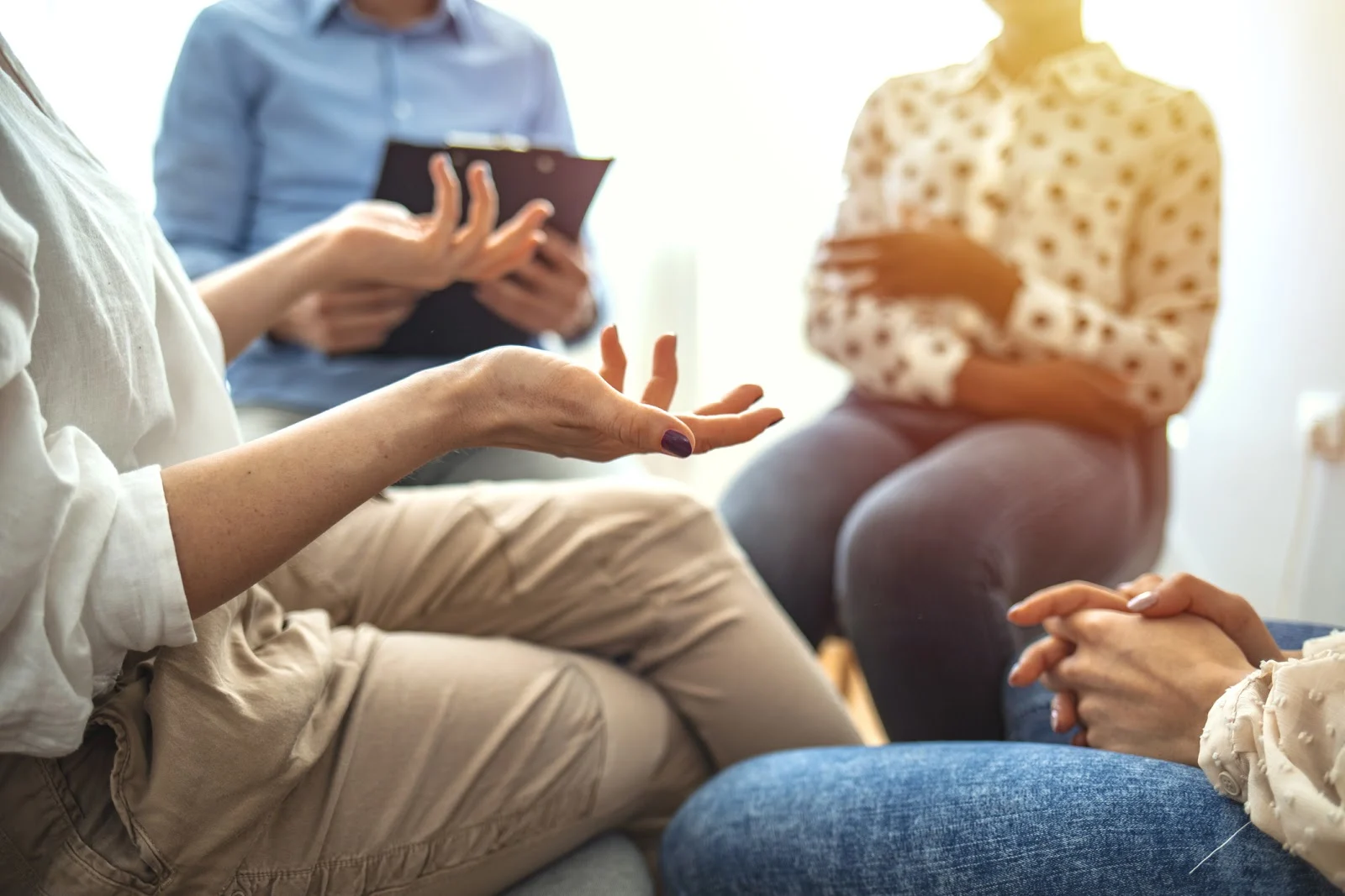 Woman gestures while discussing something difficult during a support group or group therapy session. A mental health professional is taking notes in the