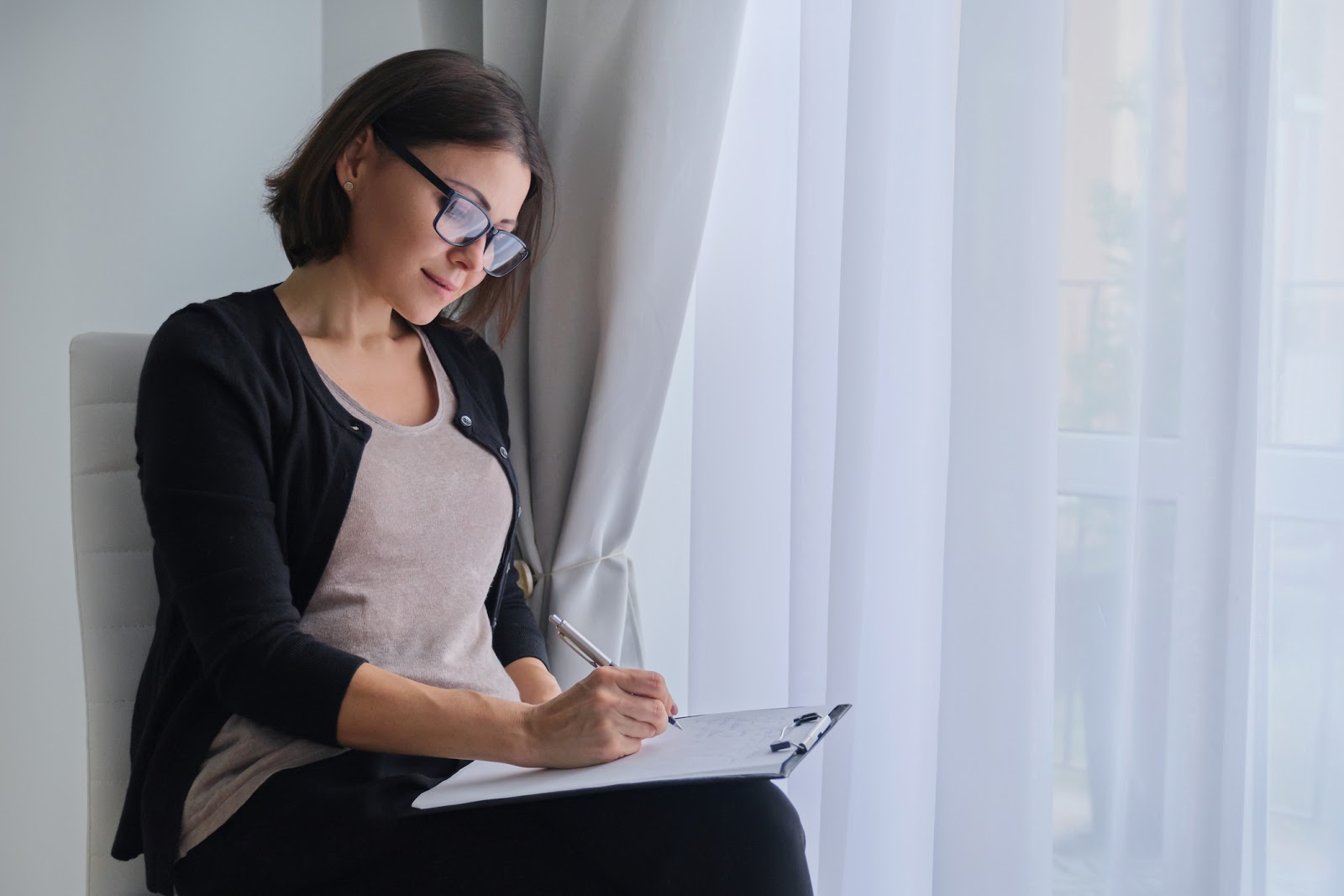 A woman with glasses sitting down filling out paperwork on a clipboard