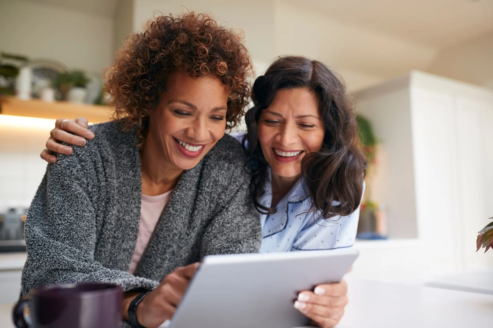 Adult female friends standing in the kitchen, smiling while looking at something on a tablet
