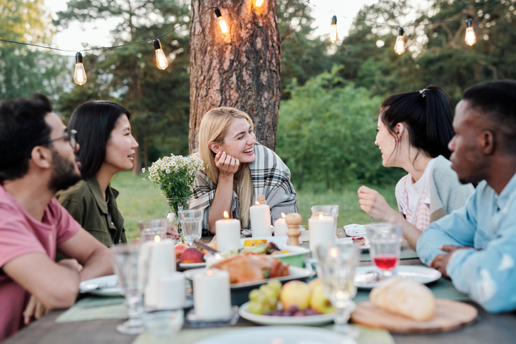 Young joyful couples sitting by served festive table under pine tree, talking, laughing and enjoying tasty food at outdoor dinner or party