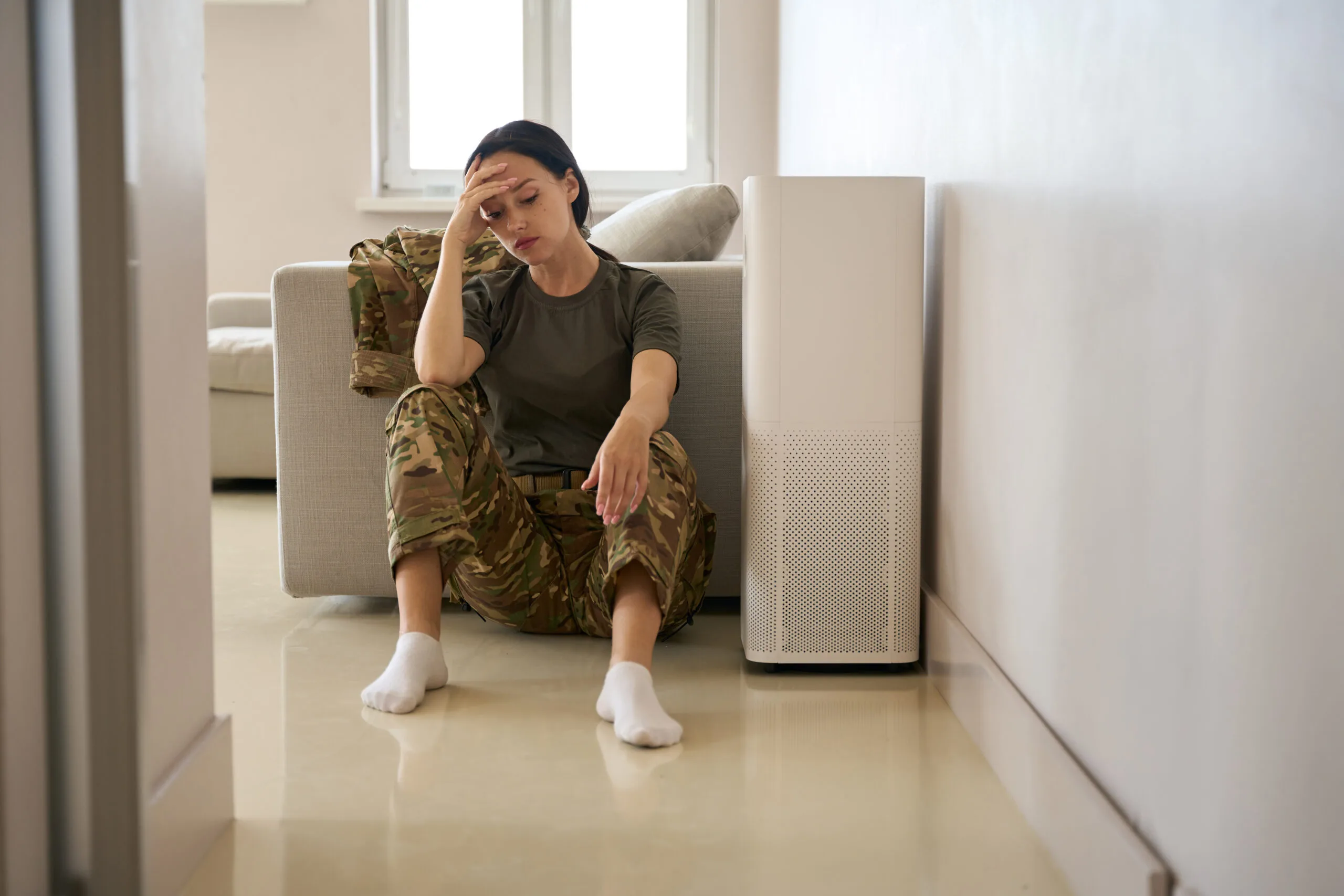 Upset young woman in camouflage clothes sits on the floor in a bright room near the sofa