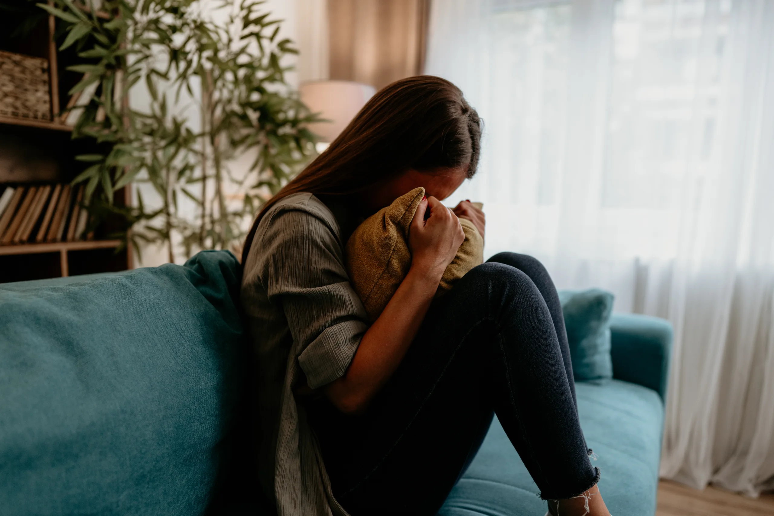 Sad young woman sitting on a sofa, hugging a pillow and grieving her past social life after becoming sober
