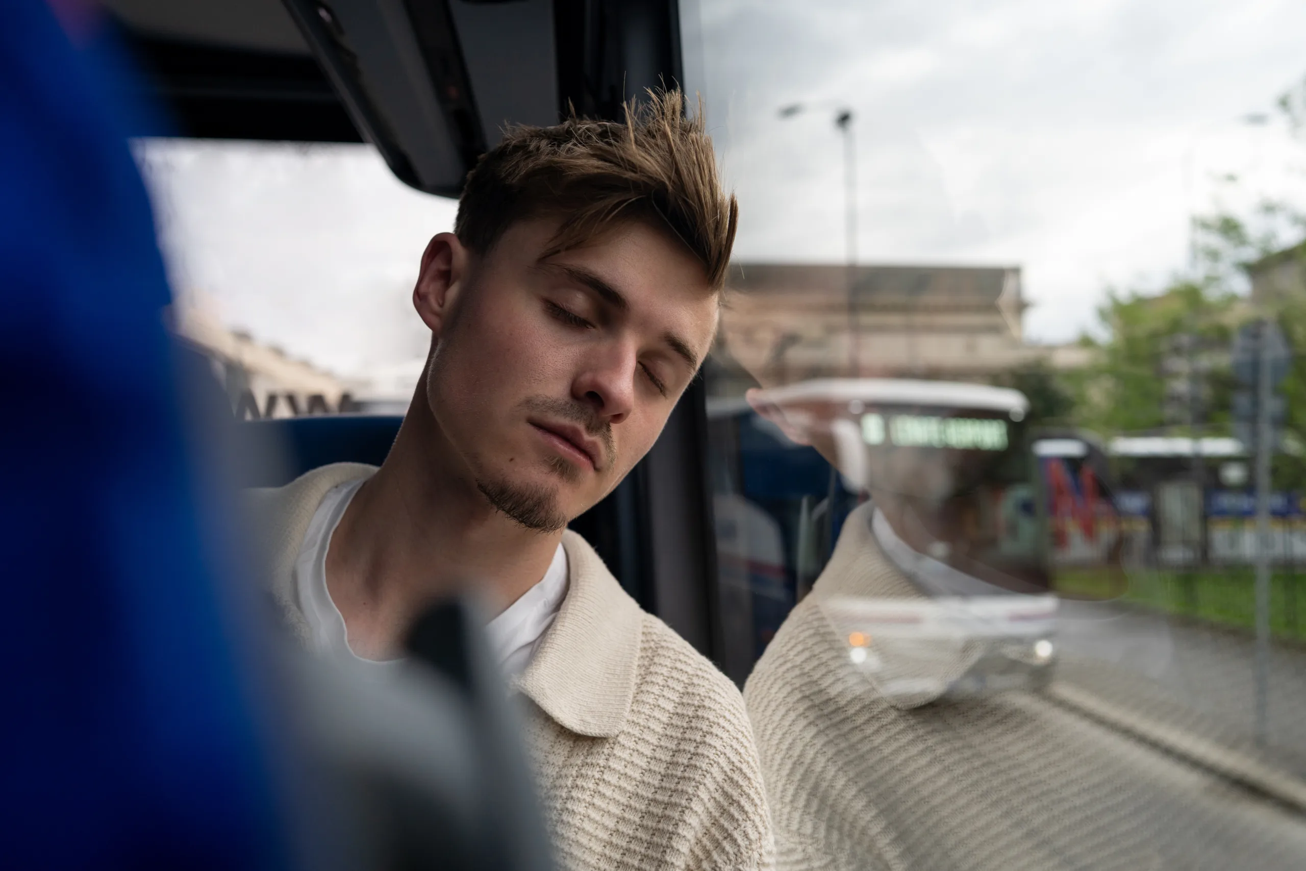 A close-up of a young adult male sleeping on a coach. His head is pressed against the window, with other coaches and city life outside in the background. Located in Milan, Italy