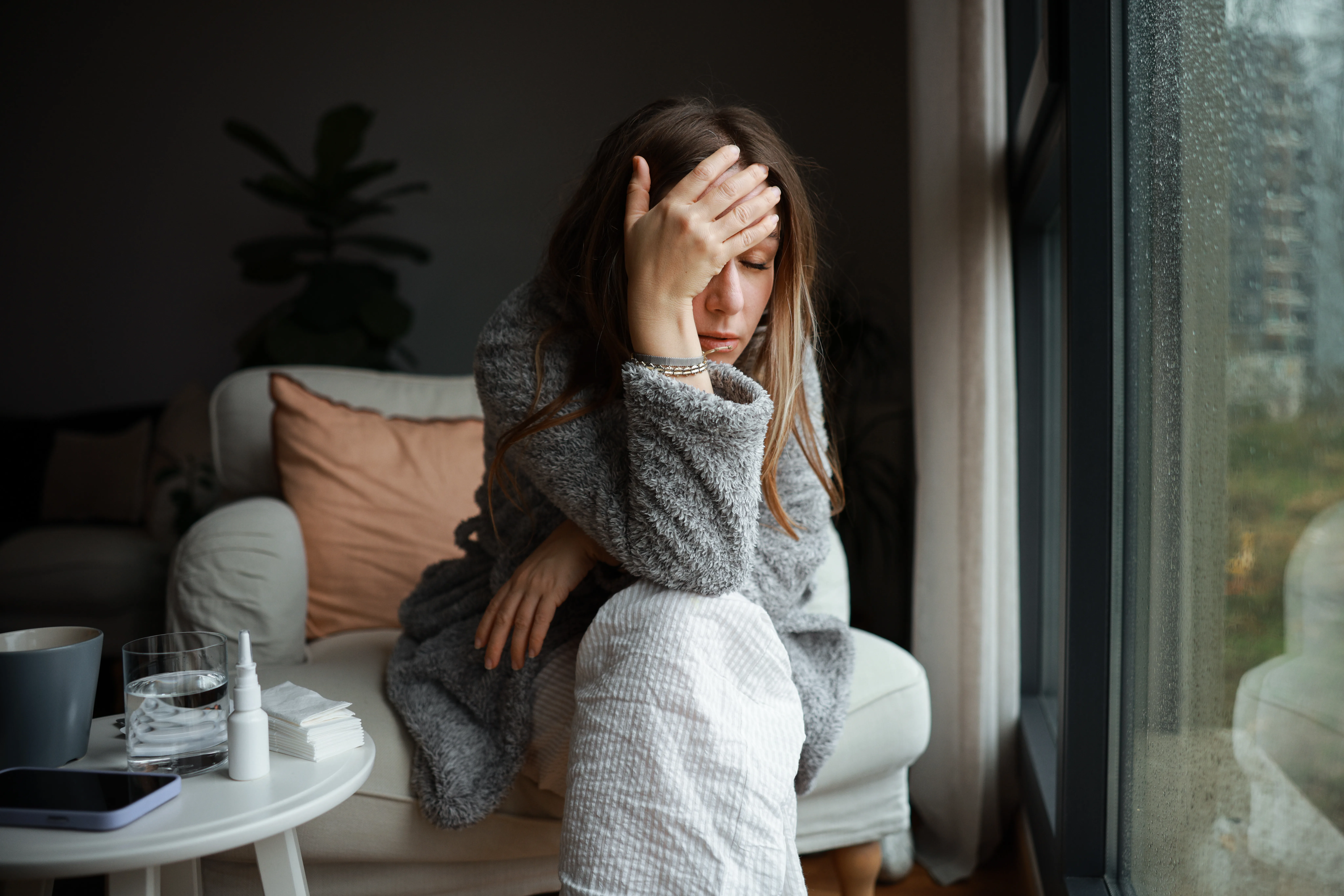 Young woman suffering from strong headache or migraine sitting with glass of water in the kitchen, millennial guy feeling intoxication and pain touching aching head, morning after hangover concept
